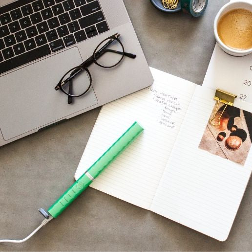 Notebook, glasses, and a green toothbrush plugged into a laptop on a desk with a cup of coffee.