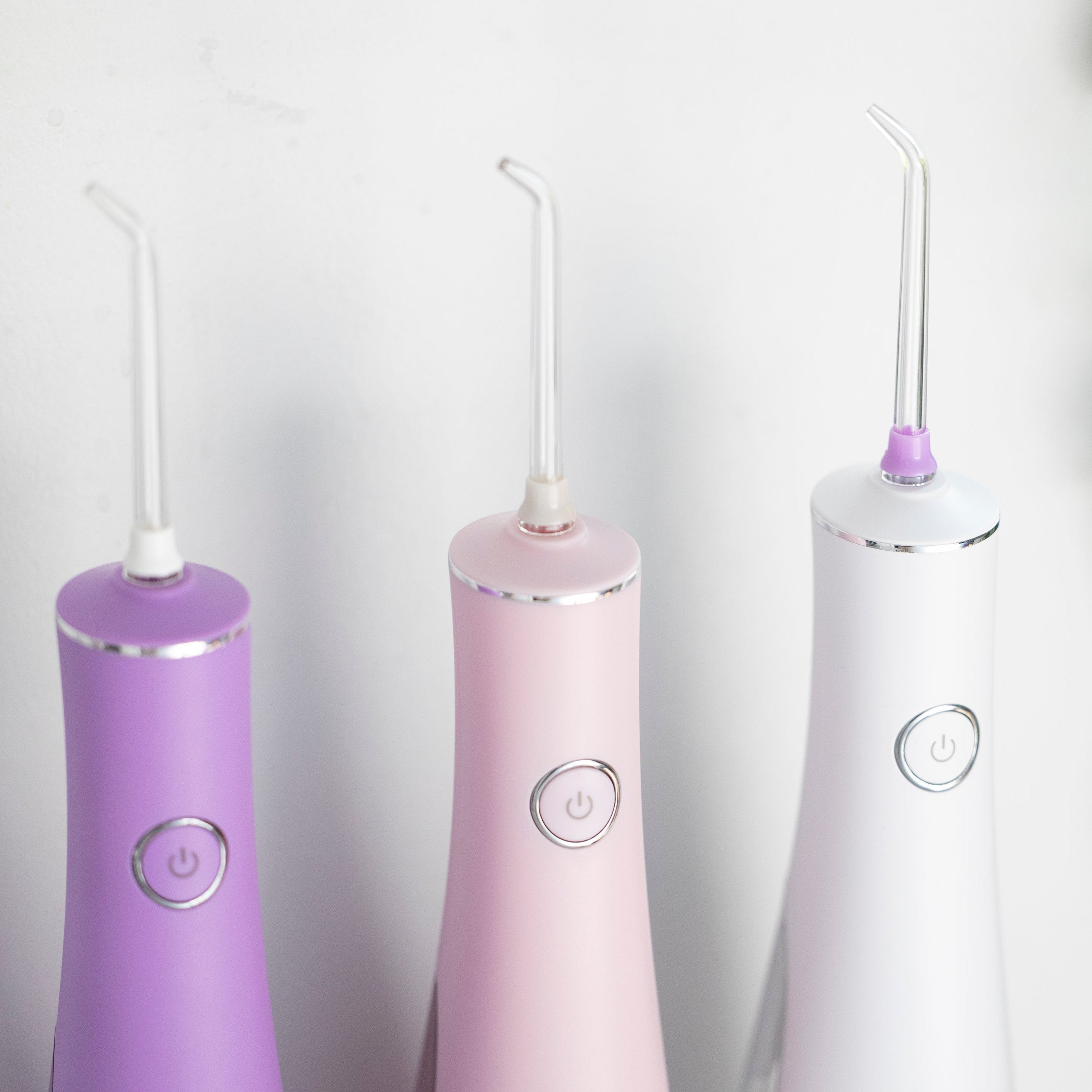 Three dental water flossers in purple, pink, and white on a light gray background.