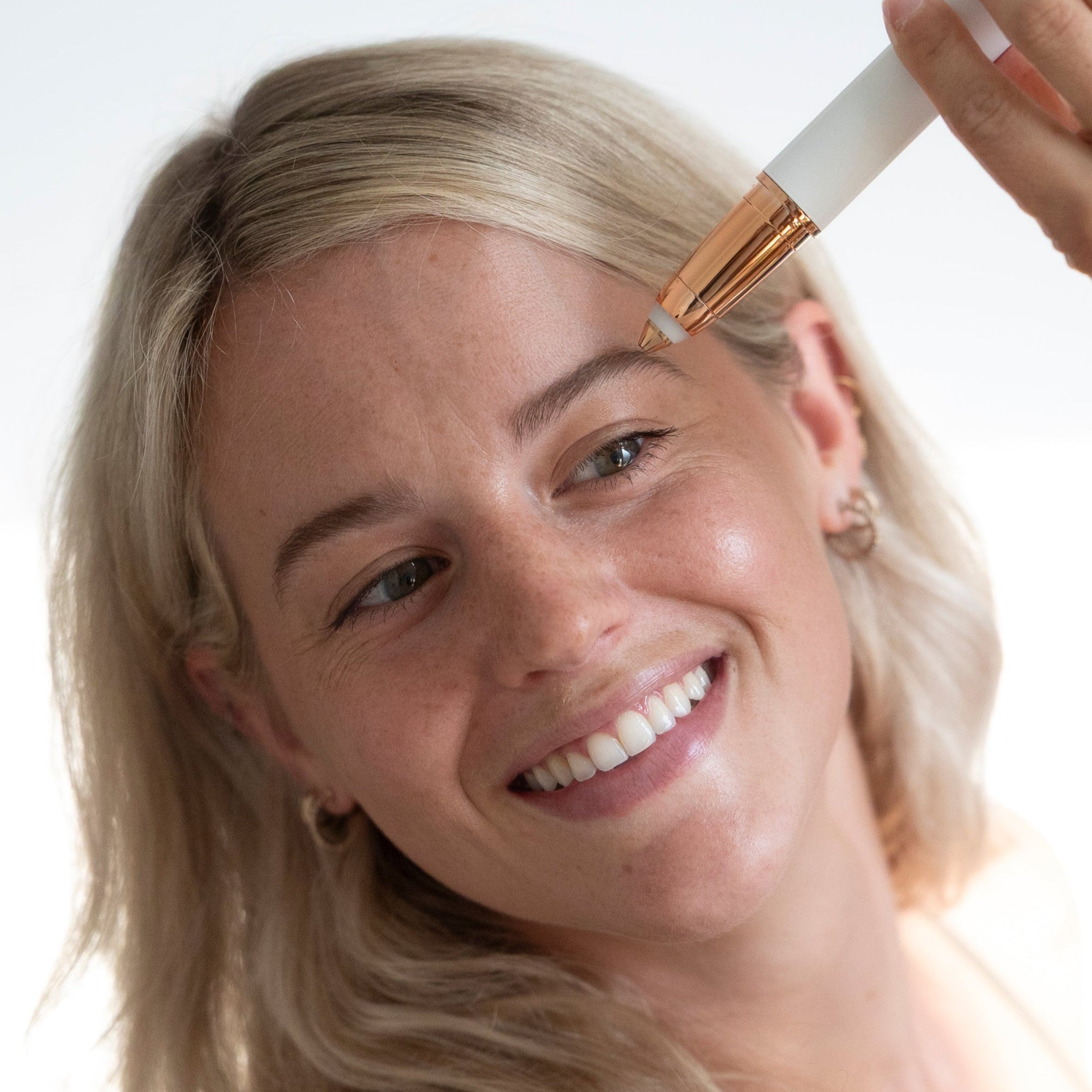 A smiling woman with blonde hair holding the white eyebrow shaper up to her eyebrow