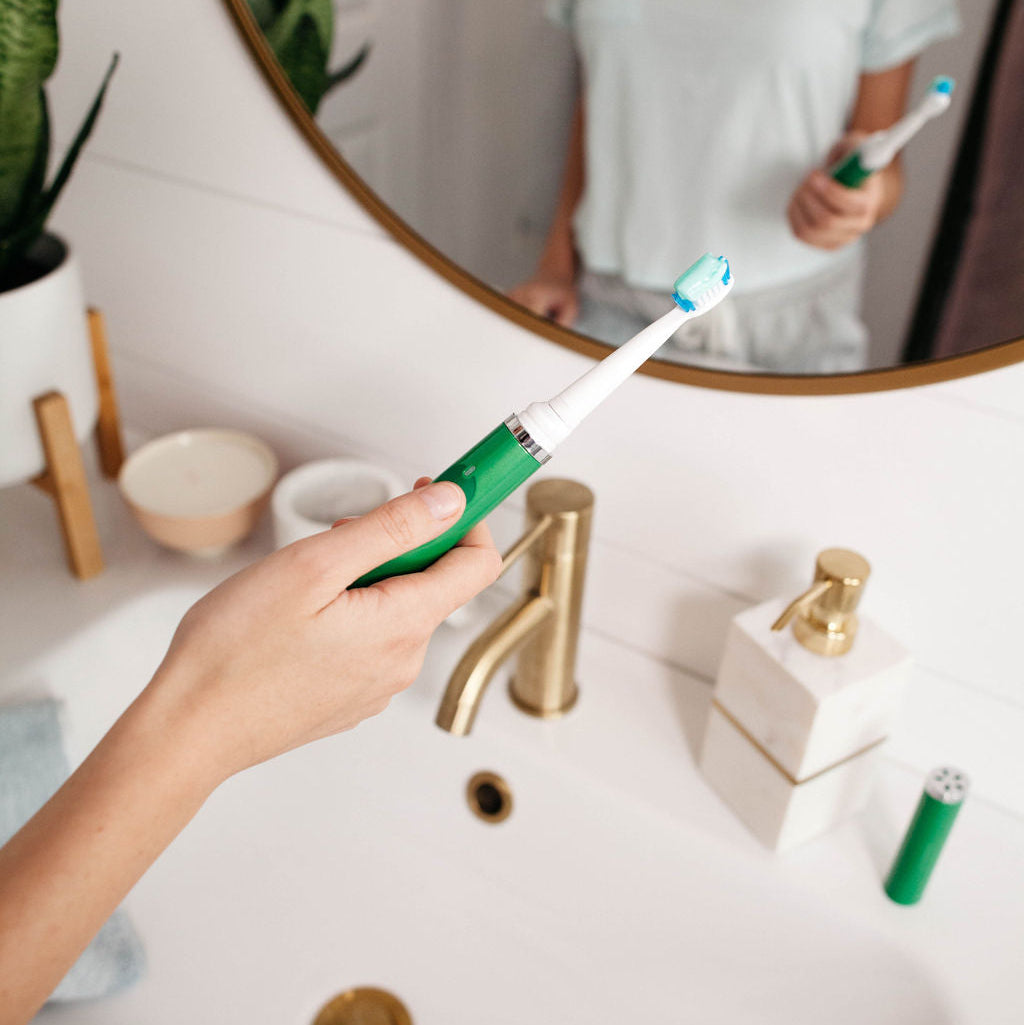 Person holding a green electric toothbrush in front of a mirror in a bathroom.