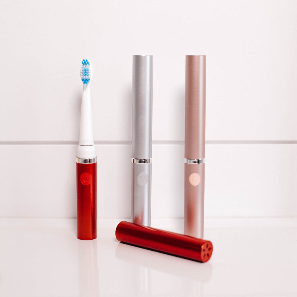 Red, silver, and rose gold electric toothbrushes on a white tiled background