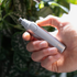 Hand holding a silver ear and nose hair trimmer against a blurred green leafy background