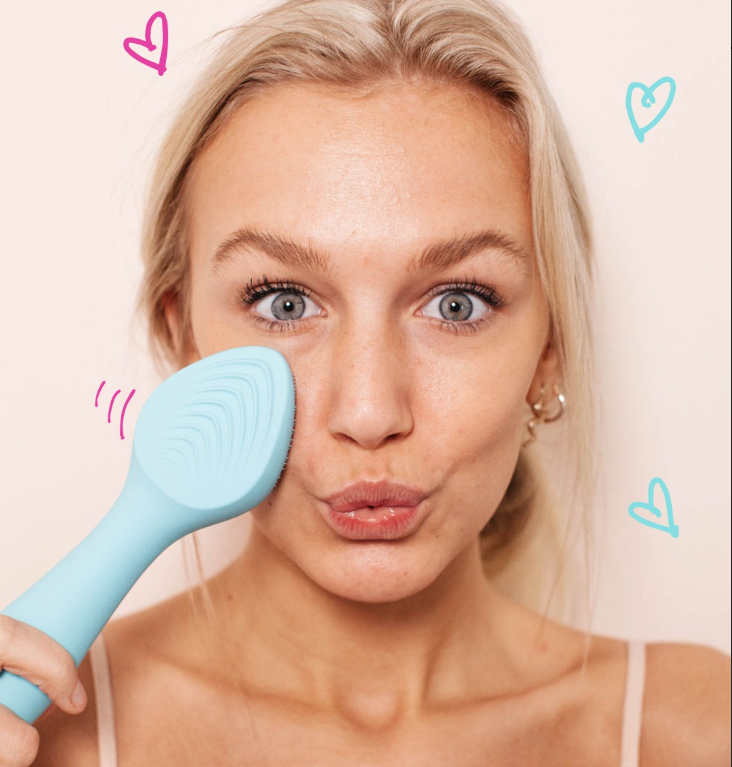 Girl with blonde hair and a pink background holding a blue facial cleansing tool to her face